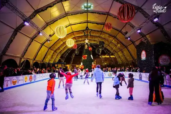 Famílias a patinar numa pista de gelo coberta, em tenda decorada com árvore de Natal e bolas iluminadas