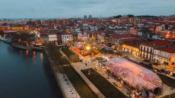 Vista aérea ao anoitecer de um recinto de Natal iluminado à beira-rio, com tenda e atrações