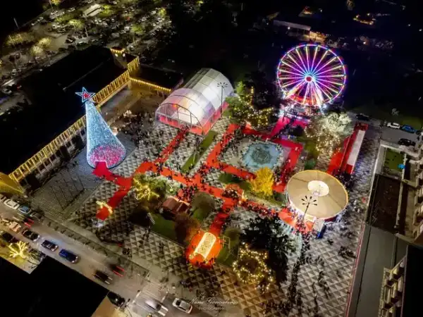 Vista aérea noturna de praça com roda gigante iluminada, árvore de Natal cónica e mercadinhos